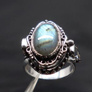 Op de afbeelding: Een zilveren ring met een blauwe en groene ovale labradorit-edelsteen gezet in een filigraan ontwerp. De ring heeft een scharnierende opening aan de zijkant.