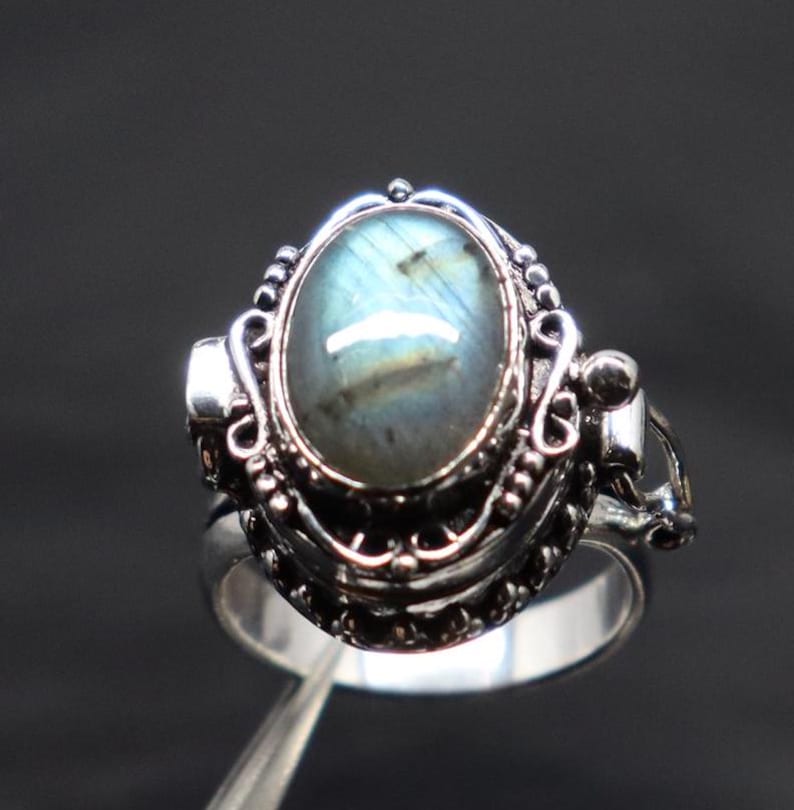 Op de afbeelding: Een zilveren ring met een blauwe en groene ovale labradorit-edelsteen gezet in een filigraan ontwerp. De ring heeft een scharnierende opening aan de zijkant.