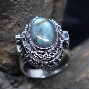 Op de afbeelding: Een zilveren ring met een scharnierend compartiment en een blauwe labradorit edelsteen. De ring is versierd met ingewikkelde bloemmotieven.