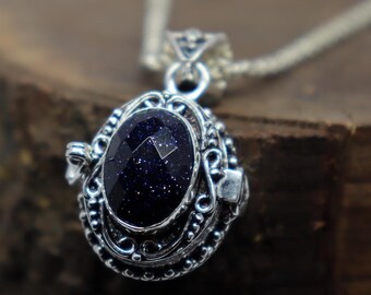 Puede incluir: Un collar con colgante plateado con una piedra preciosa ovalada de color azul oscuro. La piedra preciosa está engastada en un engaste plateado ornamentado y detallado con volutas decorativas. La cadena del collar también es plateada.