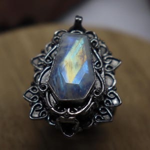 Peut inclure: Bague argentée ornée d'une grande pierre précieuse rectangulaire irisée. La pierre affiche des couleurs bleues, jaunes et vertes. La bague présente un design filigrané orné autour de la pierre, pour un style vintage.