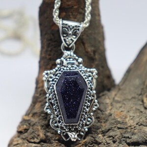 Puede incluir: Un collar con un colgante de plata con una piedra preciosa azul oscuro en forma de ata&uacute;d. El colgante est&aacute; decorado con filigrana de plata intrincada.