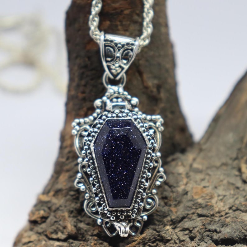 Puede incluir: Un collar con un colgante de plata con una piedra preciosa azul oscuro en forma de ata&uacute;d. El colgante est&aacute; decorado con filigrana de plata intrincada.