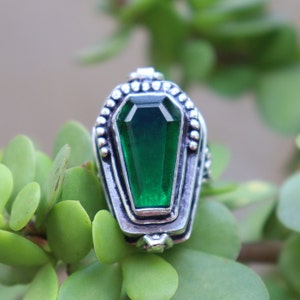 Op de afbeelding: Een zilveren ring met een groene edelsteen gezet in een kistvormig ontwerp. De ring is versierd met kleine zilveren kralen rond de rand van de kist.