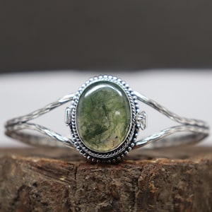 May include: Silver-toned bracelet featuring an oval moss agate gemstone. The gemstone is a mix of green and white, set in a beaded bezel. The bracelet has a textured, open design.