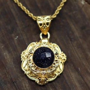 Può includere: Una collana con pendente dorato con una pietra blu scuro sfaccettata. Il pendente ha un design ornato a filigrana ed è sospeso a una catena dorata abbinata. La collana è esposta su una superficie di legno.