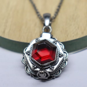 May include: A silver locket necklace with a red gemstone pendant. The locket has a detailed design with a filigree border and a small clasp.