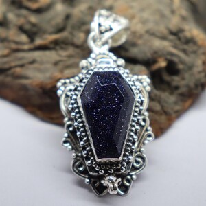 Puede incluir: Un colgante de plata en forma de ata&uacute;d con una piedra preciosa azul oscuro engastada en el centro. La piedra preciosa tiene una textura brillante y moteada.