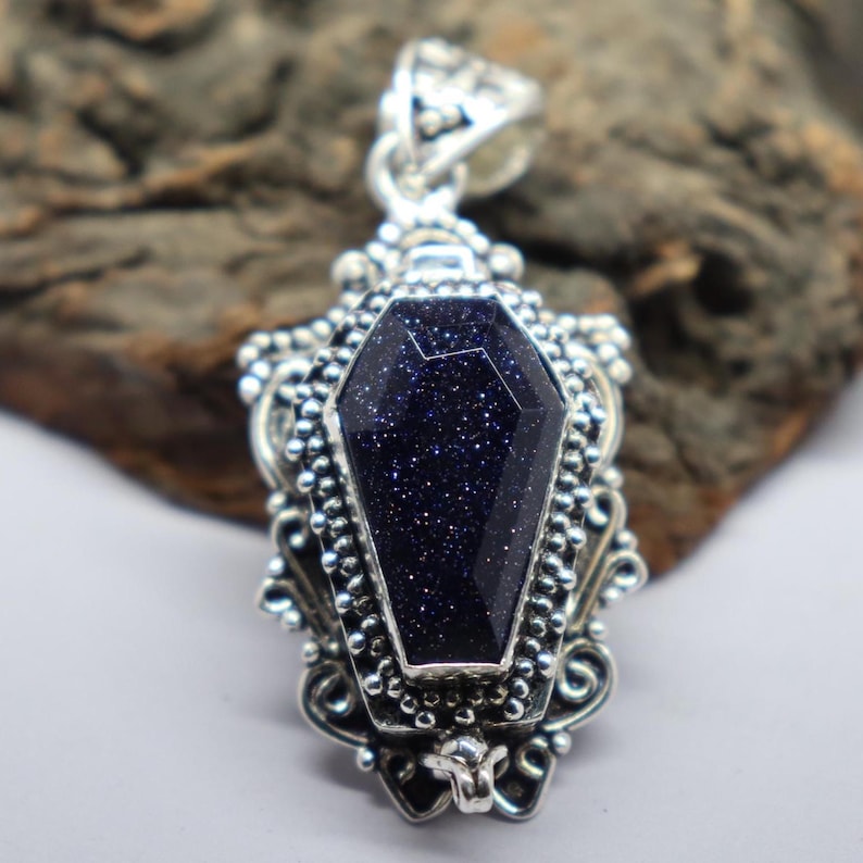 Puede incluir: Un colgante de plata en forma de ata&uacute;d con una piedra preciosa azul oscuro engastada en el centro. La piedra preciosa tiene una textura brillante y moteada.