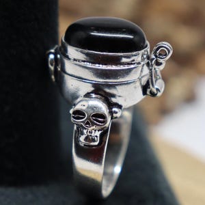 Peut inclure: Une bague en argent avec une pierre noire sertie dans une bande de bague en forme de crâne. La bague est une boîte à charnière avec une petite chaîne attachée au couvercle.