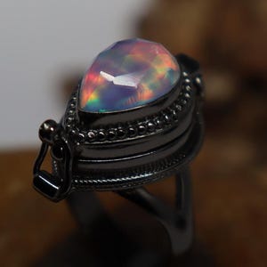 May include: A black ring with a hinged locket featuring a pear-shaped iridescent opal gemstone.