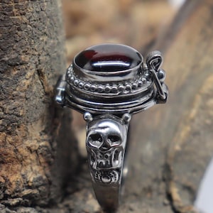 Puede incluir: Un anillo de plata con un diseño de calavera en la banda y un compartimento con bisagras con una piedra preciosa roja en la parte superior.