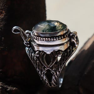 Puede incluir: Un anillo de plata con un compartimento con bisagras y una piedra de &aacute;gata verde musgo. El anillo tiene un dise&ntilde;o intrincado con un patr&oacute;n de filigrana.