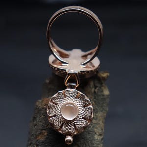 May include: A rose gold ring with a dangling charm. The charm is a round, silver-toned disc with a delicate floral design and a clear, round gemstone in the center.