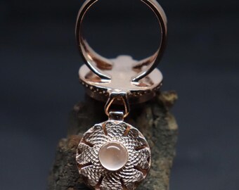 Puede incluir: Un anillo de oro rosa con un colgante. El colgante es un disco redondo plateado con un delicado diseño floral y una piedra redonda transparente en el centro.