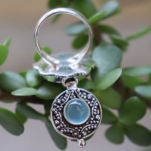 Pode incluir: Um anel de prata com um pingente de pedra azul. O anel tem um design intrincado e o pingente é redondo com uma borda detalhada.