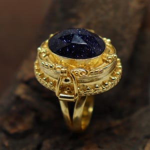 May include: A gold ring with a large, oval, dark blue gemstone with sparkling inclusions. The ring has detailed embellishments around the stone and on the band, with a decorative clasp feature.