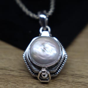 Op de afbeelding: Een zilveren ketting met een zeshoekige hanger met een grote, witte parelmoer inleg. De hanger heeft een scharnierend deksel en ingewikkelde details rond de randen.