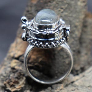 Op de afbeelding: Een zilveren ring met een scharnierend compartiment en een heldere ovale edelsteen. De ring is versierd met ingewikkelde details en heeft een gladde, gepolijste afwerking.