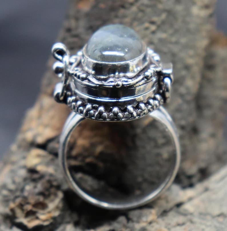 Op de afbeelding: Een zilveren ring met een scharnierend compartiment en een heldere ovale edelsteen. De ring is versierd met ingewikkelde details en heeft een gladde, gepolijste afwerking.