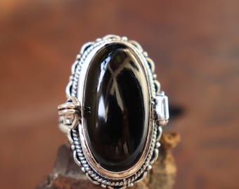 Könnte beinhalten: Ein silberner Ring mit einem ovalen schwarzen Onyxstein, der in einer detaillierten silbernen Fassung sitzt. Der Ring hat ein dekoratives Design um den Stein herum.