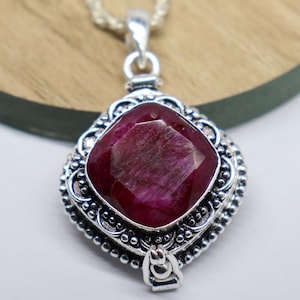 May include: A silver pendant necklace featuring a large, faceted ruby gemstone. The ruby is a deep red color and is set in an ornate, detailed silver setting. The pendant hangs from a delicate silver chain.