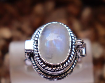 Puede incluir: Un anillo de plata con un cabujón ovalado de piedra lunar engastado en un engaste de plata detallado. El anillo tiene una abertura con bisagra en el lateral.