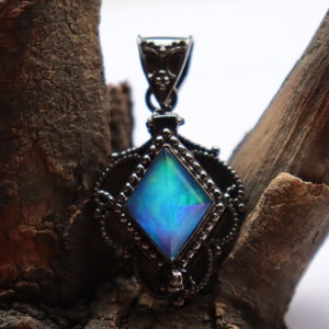 Puede incluir: Un colgante de plata con una piedra preciosa azul iridiscente engastada en un marco de forma de diamante. El colgante tiene un diseño detallado con pequeñas cuentas alrededor del borde.