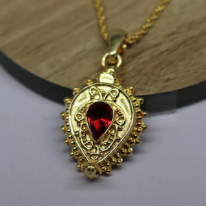 May include: A gold pendant necklace featuring a teardrop-shaped, red gemstone. The pendant has an intricate design with a beaded edge and is suspended from a fine gold chain. The necklace is presented on a wooden surface.