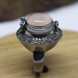 May include: A silver ring featuring a large, oval, light pink gemstone. The ring has a detailed, decorative setting with a rounded top and a wide band. The gemstone is the focal point, set in a bezel setting.