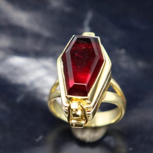 Può includere: Un anello d'oro con una grande pietra preziosa rossa sfaccettata incastonata in una montatura a forma di bara.