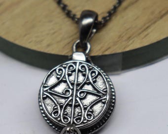 Puede incluir: Un collar con colgante redondo plateado con un diseño ornamental simétrico. El colgante presenta un patrón detallado de líneas y curvas arremolinadas dentro de un marco circular, suspendido de una cadena oscura.