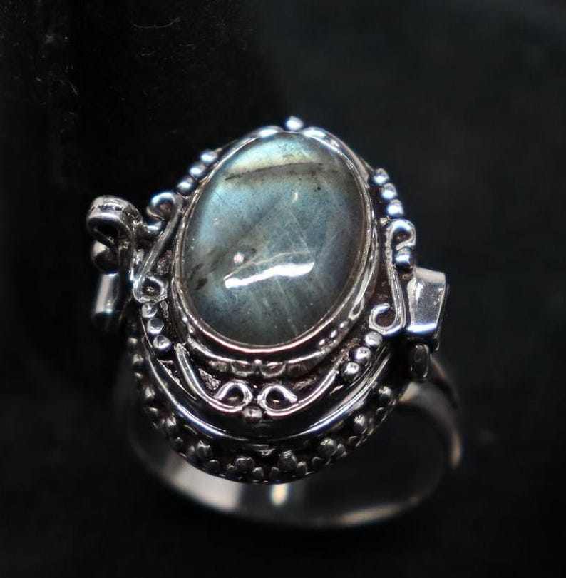 Op de afbeelding: Een zilveren ring met een scharnierend medaillon met een grote ovale labradorit-edelsteen. De ring heeft ingewikkelde zilveren details rond het medaillon.