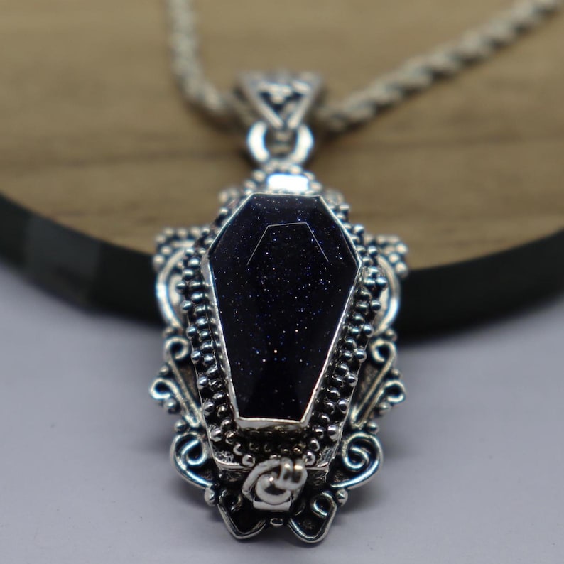 Puede incluir: Un collar de plata con un colgante de piedra negra en forma de ata&uacute;d. El colgante tiene intrincados detalles de plata.