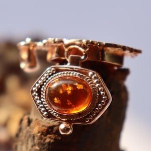 Könnte beinhalten: Ein goldener Anhänger mit einem detaillierten Design, das einen runden Bernstein in einem sechseckigen Rahmen zeigt. Der Anhänger ist an einer goldenen Kette befestigt.