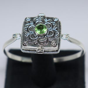 Puede incluir: Una pulsera de plata con un medallón con bisagras que presenta un intrincado diseño floral y una piedra preciosa verde en el centro.