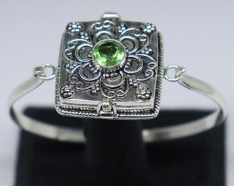 Puede incluir: Una pulsera de plata con un medallón con bisagras que presenta un intrincado diseño floral y una piedra preciosa verde en el centro.