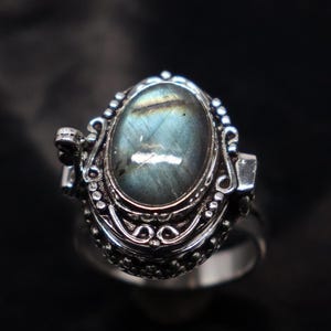 Op de afbeelding: Een zilveren ring met een scharnierend medaillon met een blauwe en groene labradorit edelsteen. Het medaillon is versierd met ingewikkelde bloemmotieven.