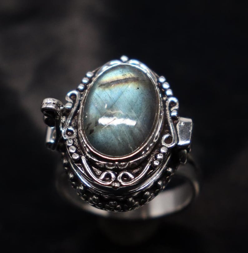 Op de afbeelding: Een zilveren ring met een scharnierend medaillon met een blauwe en groene labradorit edelsteen. Het medaillon is versierd met ingewikkelde bloemmotieven.