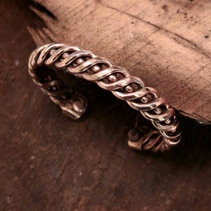 May include: A copper-colored cuff bracelet with a braided design. The bracelet features a series of interwoven, wavy strands, creating a textured surface. Small, round beads are interspersed along the design. The open-ended cuff is displayed on a wooden surface.