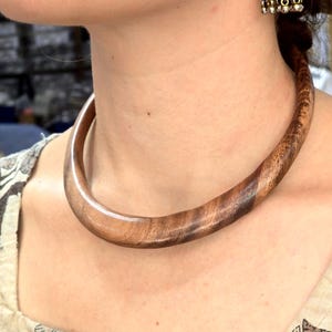 Peut inclure: Un collier torque en bois poli et lisse présentant un motif de grain de bois brun naturel. Le collier a une forme circulaire épaisse et arrondie qui repose près du cou. Une boucle d'oreille jhumka argentée avec des perles pendantes est visible.