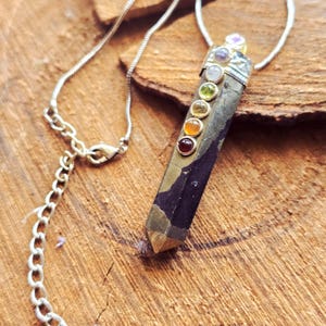 Peut inclure: Un collier à chaîne argentée avec un pendentif en pierre rectangulaire foncée orné de petites pierres précieuses colorées. Le pendentif a un capuchon en métal doré et est suspendu à une chaîne argentée délicate. Les pierres sont rouges, oranges, jaunes, vertes, blanches et violettes.