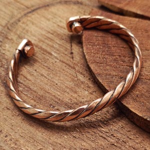 Peut inclure: Un bracelet manchette de couleur cuivre avec un motif de corde torsadée. Le bracelet a un design simple mais élégant avec une fermeture à fermoir. Le métal a une teinte brun-rougeâtre chaude.
