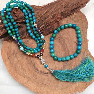 May include: A turquoise and green beaded mala necklace with a Buddha head pendant and a matching bracelet. The necklace has a long tassel. The beads are various shades of blue and green. The items are displayed on a wooden surface.