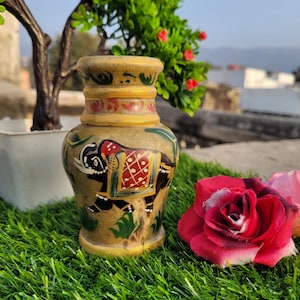 Peut inclure: Un petit vase en céramique peint à la main avec un bord doré et un motif d'éléphant. Le vase est décoré de peinture verte, rouge et noire. Il est posé sur une surface d'herbe artificielle verte.