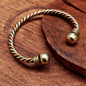 Op de afbeelding: Een metalen manchetarmband met een gedraaid ontwerp, in goud-, koper- en zilverkleuren. De armband heeft een spiraalpatroon en is afgewerkt met een kleine, ronde, goudkleurige bal aan elk uiteinde. Het ligt op een houten oppervlak.