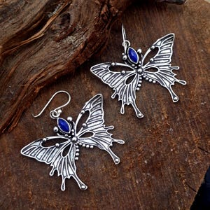 Peut inclure: Une paire de boucles d'oreilles papillon en métal argenté avec des motifs d'ailes ajourés et une pierre bleue foncée en forme de marquise au centre. Chaque boucle est suspendue à un crochet simple, avec une finition métallique détaillée.