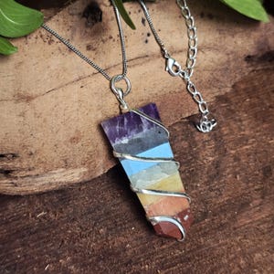 Peut inclure: Un collier avec une chaîne argentée et un pendentif rectangulaire. Le pendentif est un cristal multicouche dans les tons violets, bleus, jaunes et rouges, enveloppé de fil d'argent. Le collier est présenté sur une surface en bois.