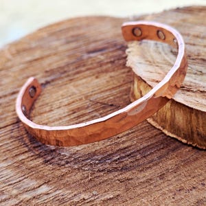 Peut inclure: Un bracelet manchette en cuivre martelé. Le bracelet ouvert a une surface texturée et une couleur cuivrée chaude. Le bracelet est présenté sur une surface en bois rustique. Le bracelet a deux petits aimants à chaque extrémité.