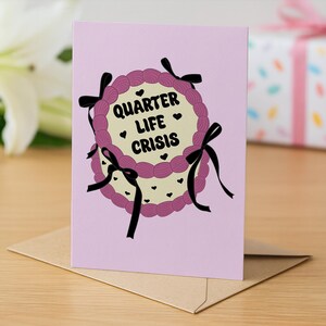 May include: A greeting card with a pink background and a cake design. The cake has a cream-colored center with the words "Quarter Life Crisis" and small black hearts. The cake is decorated with purple frosting and black bows.